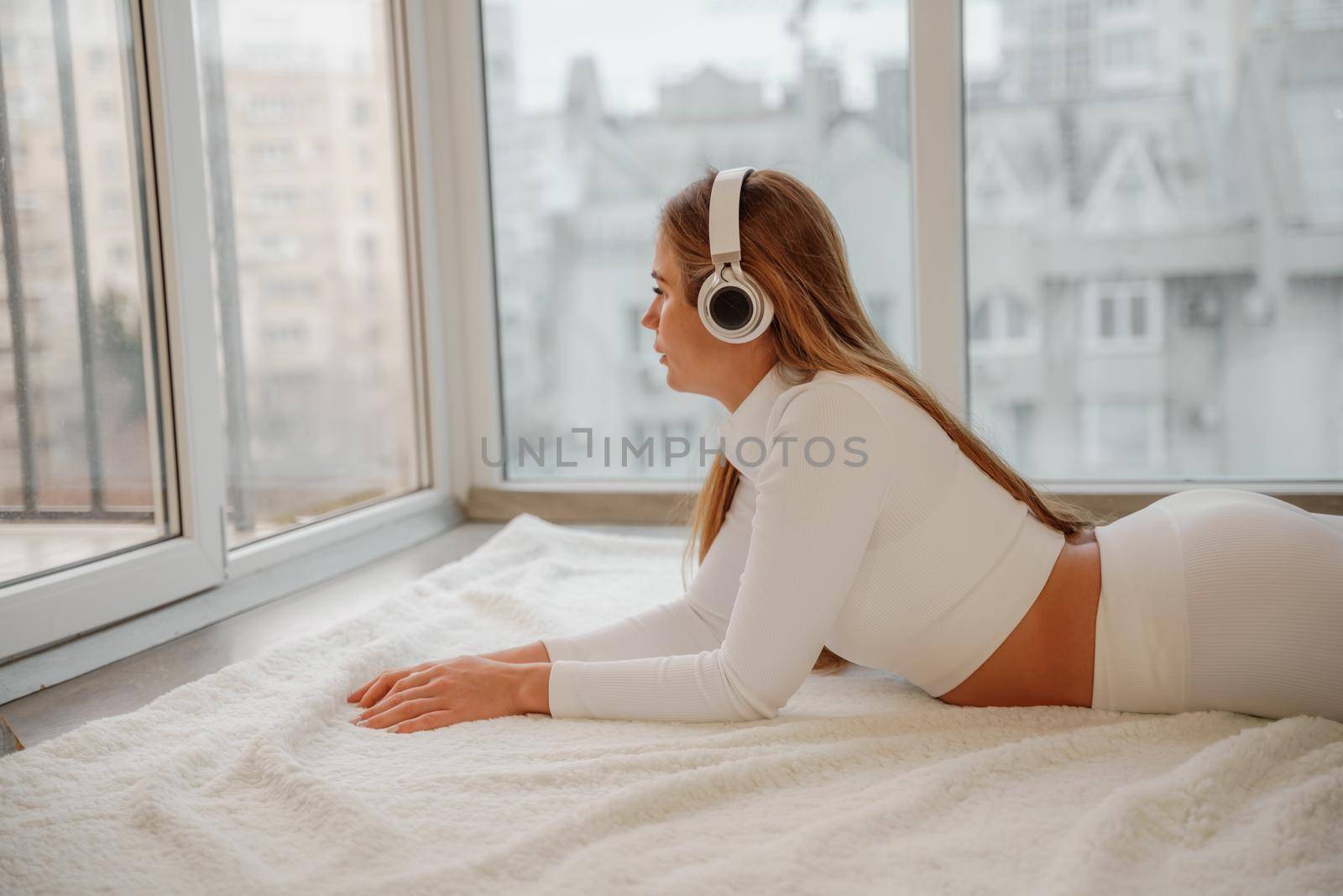 Side view portrait of relaxed woman listening to music with headphones lying on carpet at home. She is dressed in a white tracksuit. by Matiunina