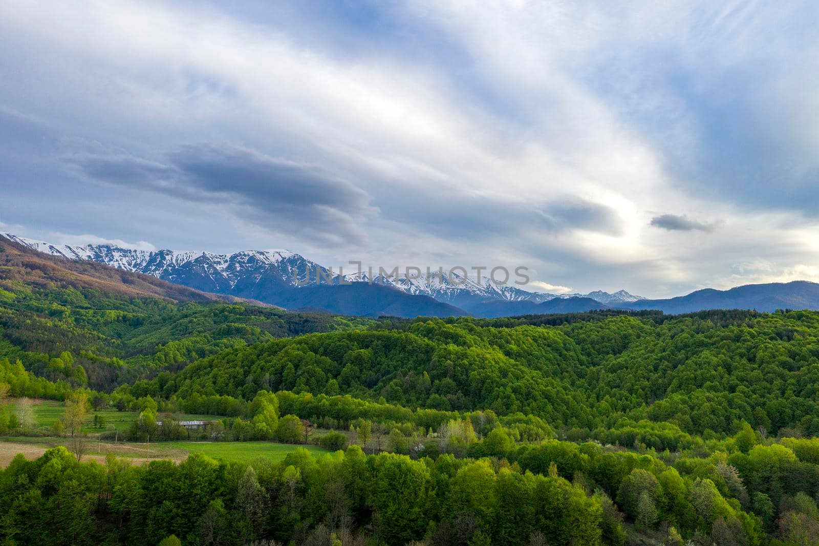 Sunny mountain scenery with vivid green forest on hill and snowy mountains in sunlight in low clouds. by EdVal