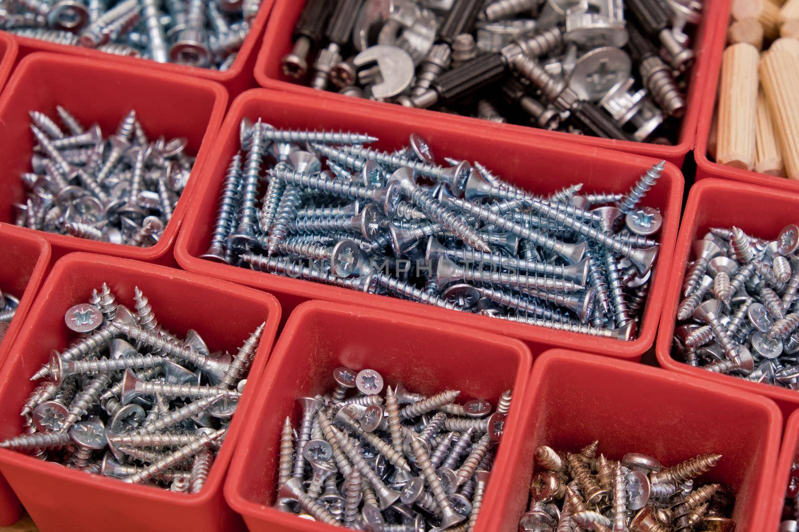 Various size and shape of screws, bolts, wooden pegs selection in red plastic tray box on a wooden background. Assembly kit for furniture. Selective focus.