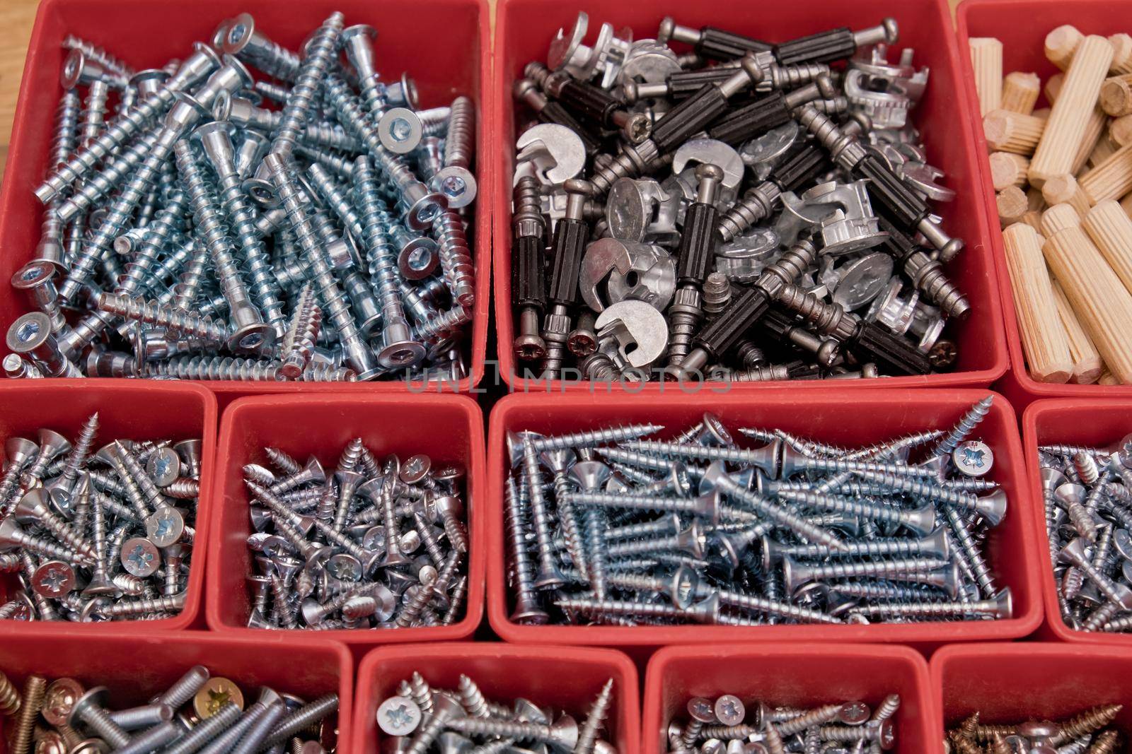 Various size and shape of screws, bolts, wooden pegs selection in red plastic tray box on a wooden background. Assembly kit for furniture. Selective focus.