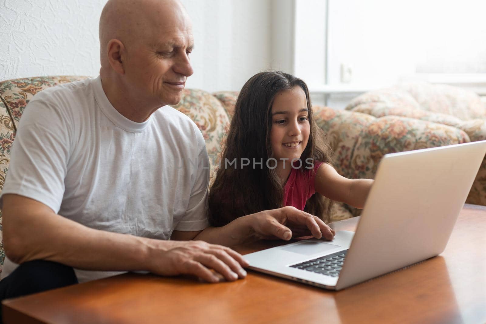 Happy little girl hugging smiling grandfather sitting on sofa with laptop