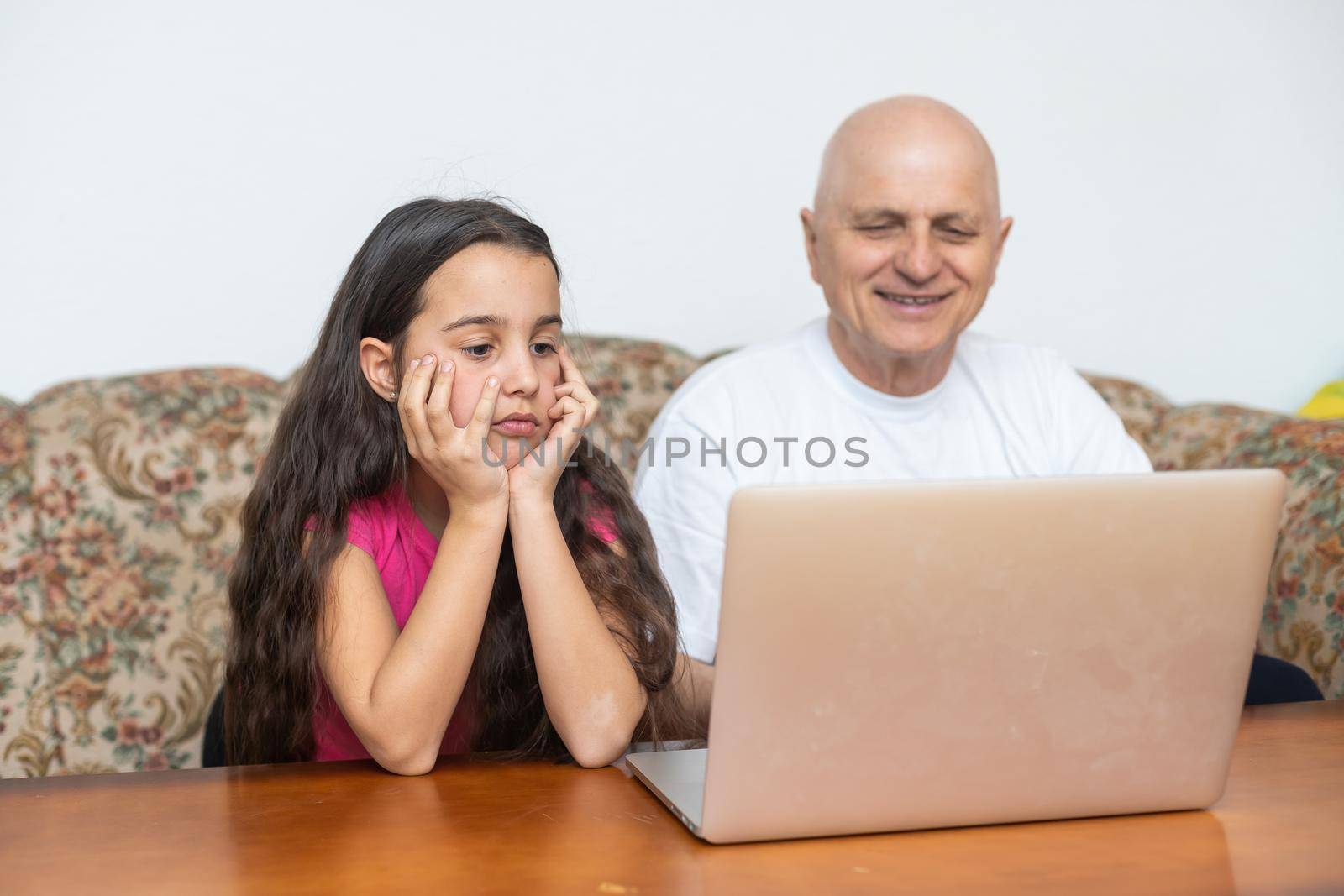 Adorable little girl hugging happy grandfather using laptop at home