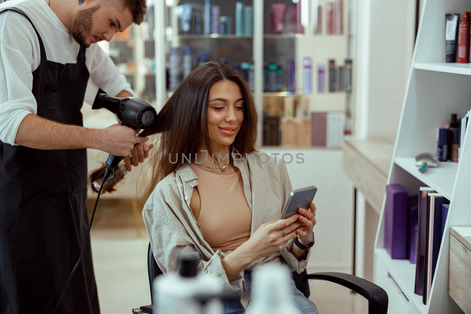 Beauty salon female customer using her phone while hairstylist doing her hair. Hair care, hairstyle