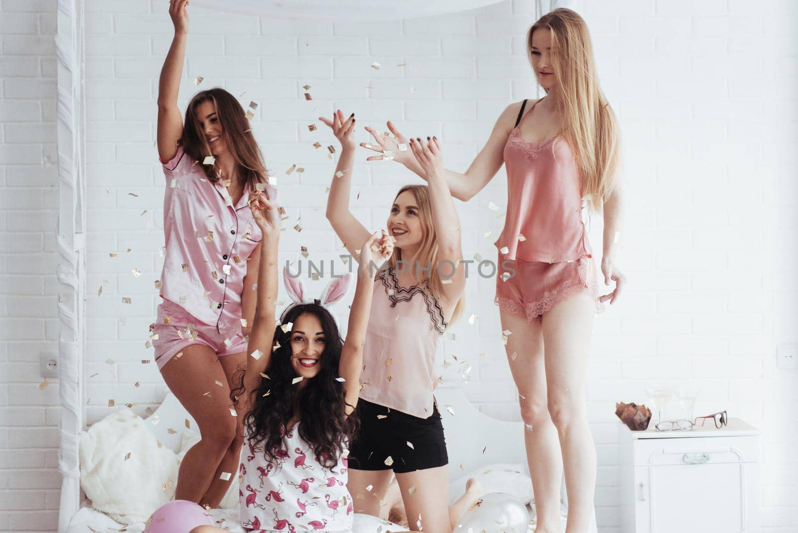 Hands up. Confetti in the air. Young girls have fun on the white bed in nice room.