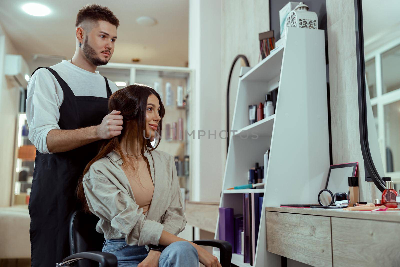 Beautiful woman with long hair looking pleased while hairstylist doing her hair at beauty salon by Yaroslav_astakhov