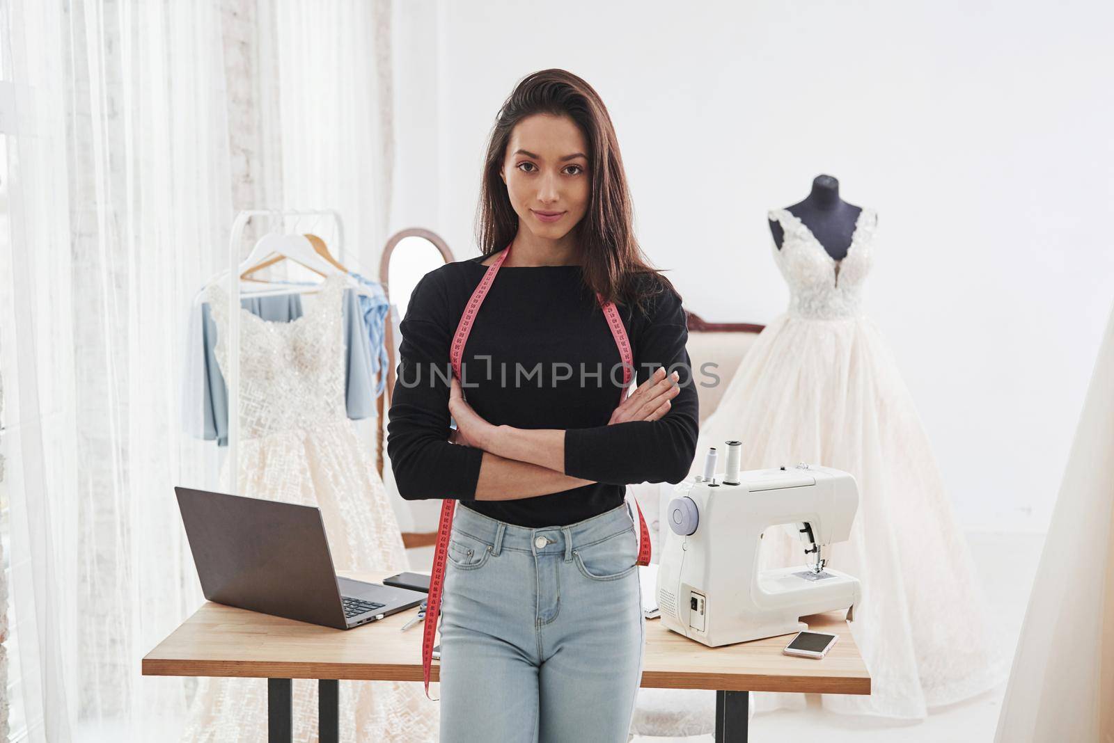 Looking into the camera with arms crossed. Portrait of female fashion designer standing in the workshop and table with instruments behind.