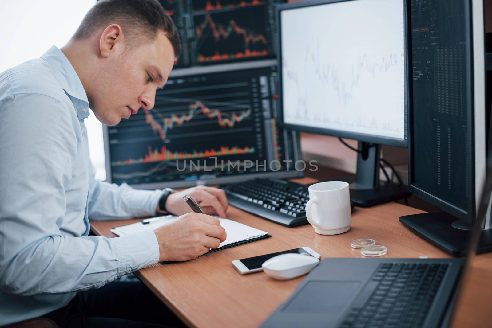 Writing on the notepad. Man working online in the office with multiple computer screens in index charts.