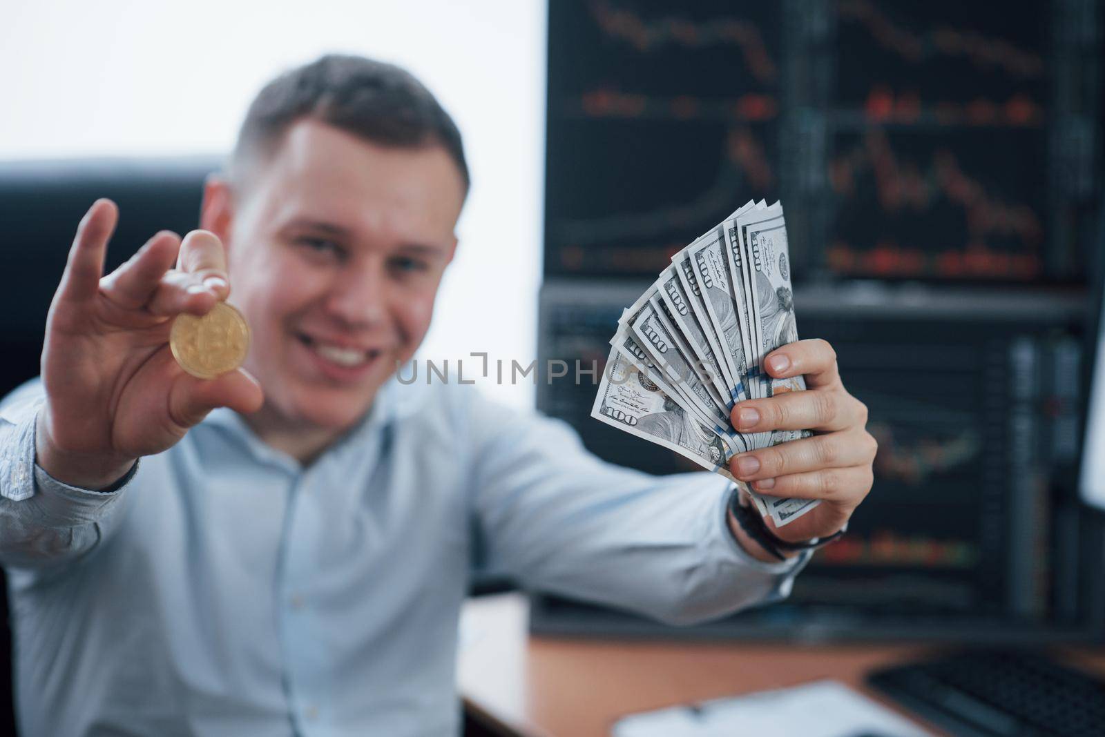 Happy rich man. Businessman holding bitcoin and money in hands while sitting in modern office with many monitors with graphs.