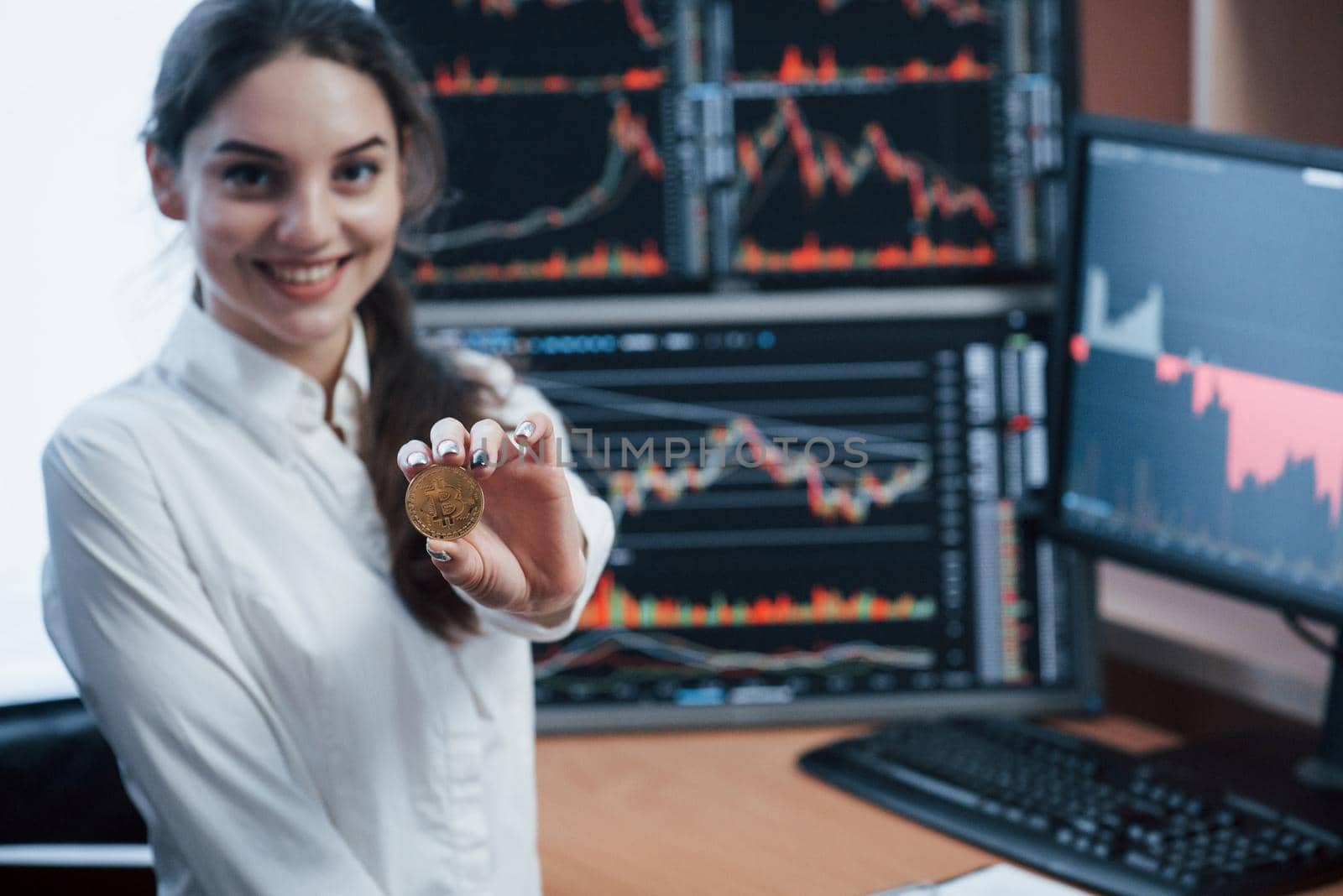 Nice daylight. Beautiful woman holding bitcoin in hands while standing in modern office with many monitors with graphs.
