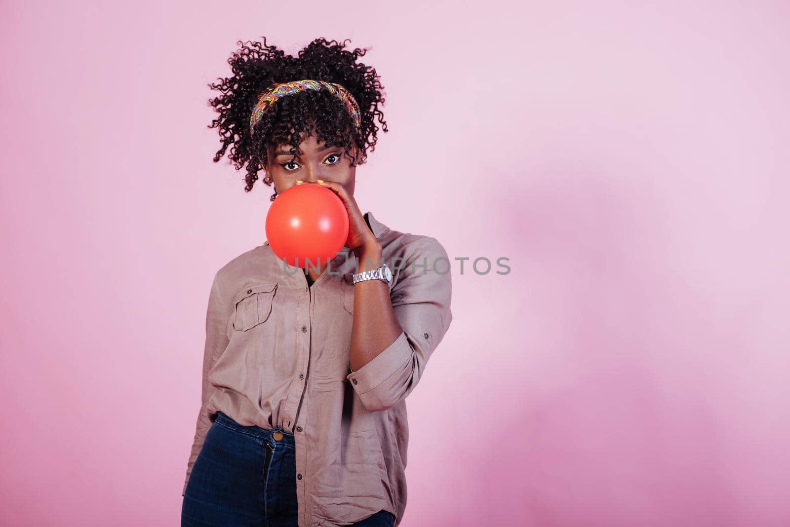 Casual clothes. Portrait of young afro american girl blowing red balloon at pink studio background.