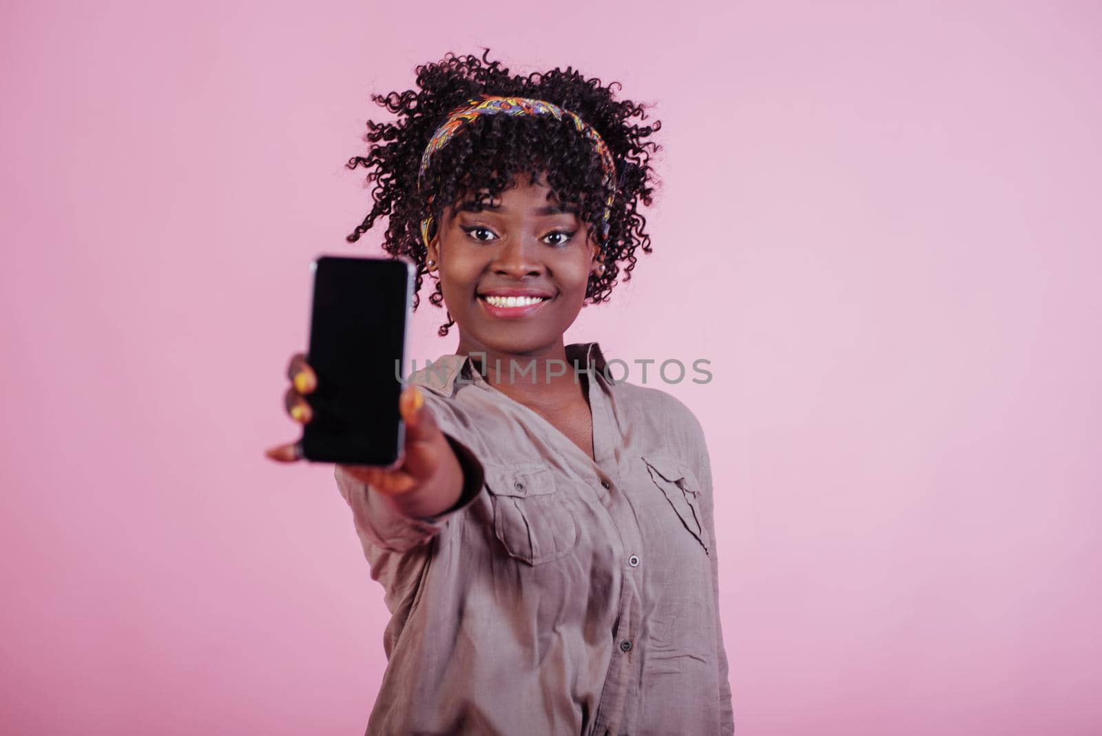 Holding black phone in hand. Attractive afro american woman in casual clothes at pink background in the studio.