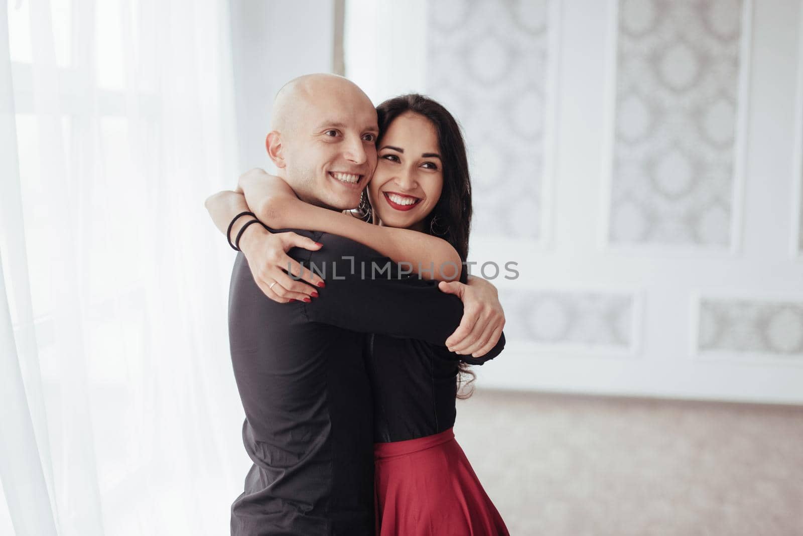 Cute and cheerful people. Hug each other and smiling. Portrait of happy couple indoors. Bald guy and brunette woman stands in the white room.