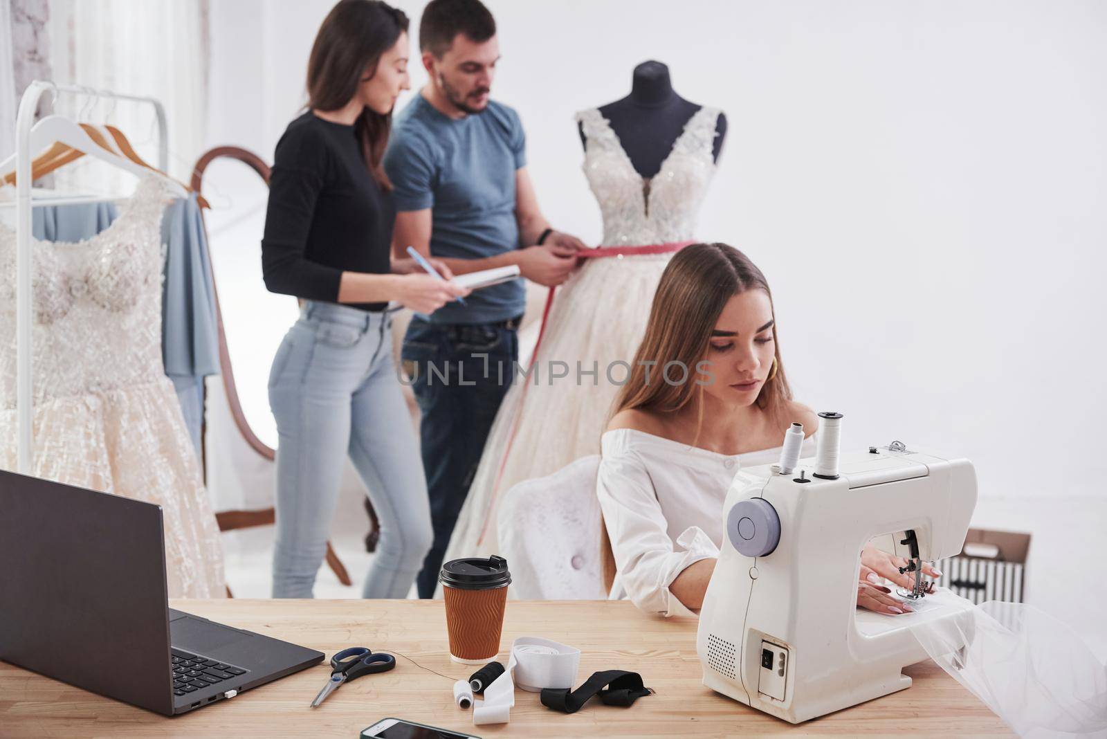 Girl and man measures dress. Female fashion designer works on the new clothes in the workshop with group of people behind.