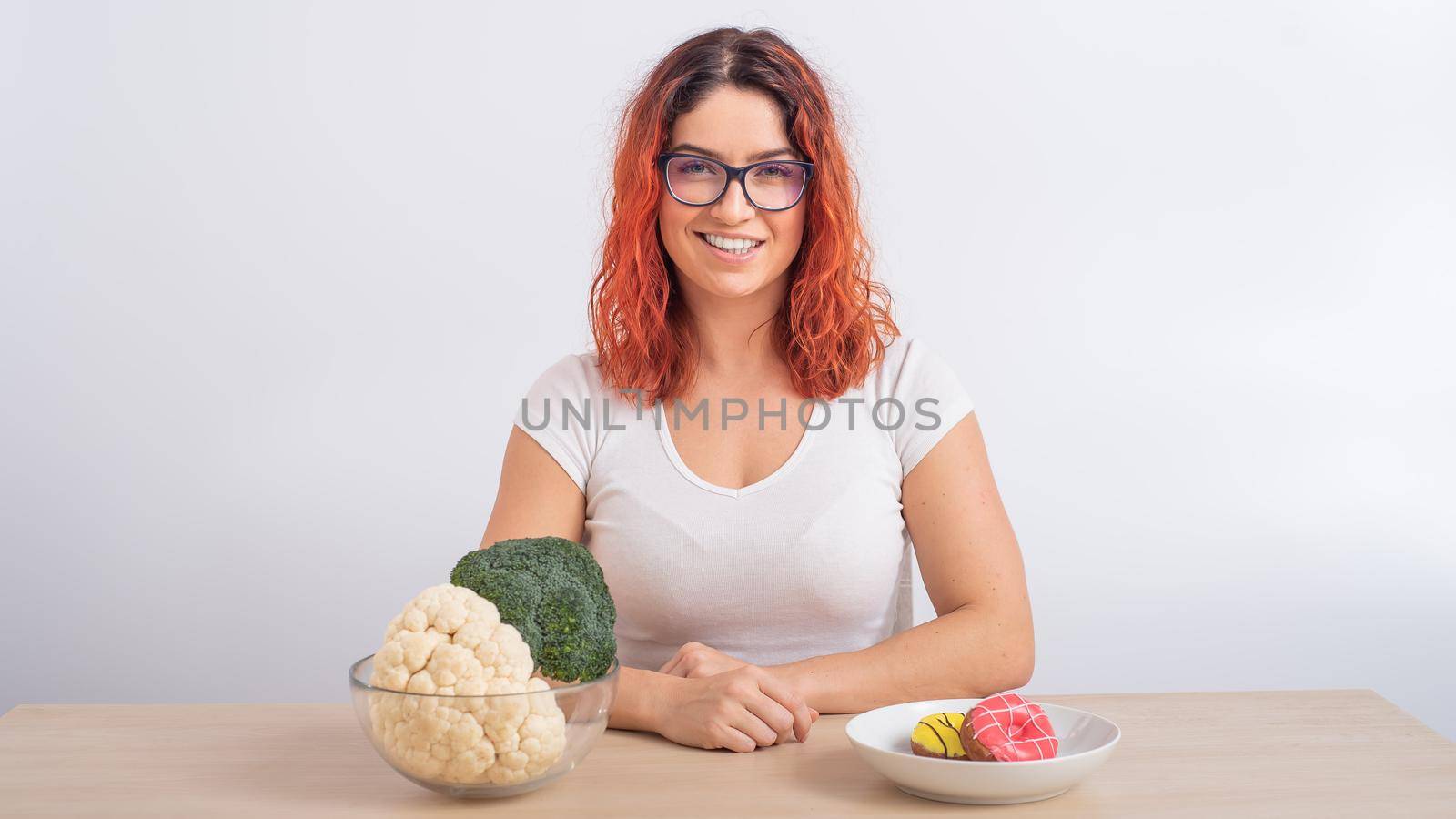 Caucasian woman prefers healthy food. Redhead girl chooses between broccoli and donuts on white background
