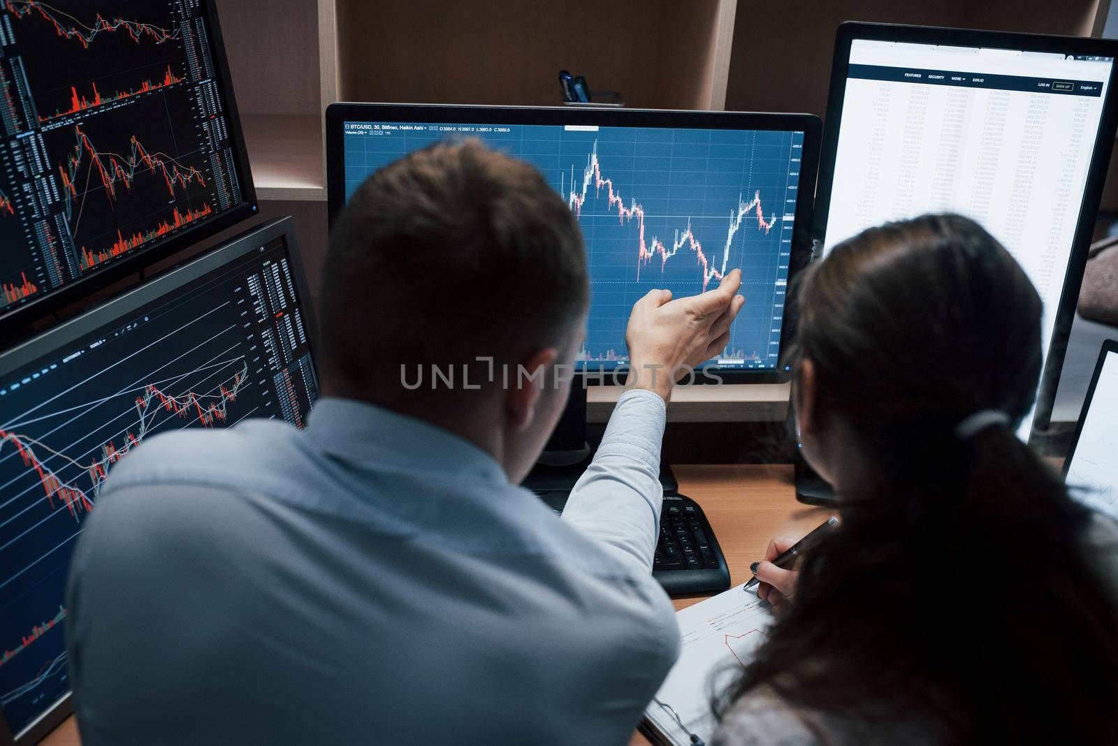 View from the back. Team of stockbrokers are having a conversation in a office with multiple display screens.