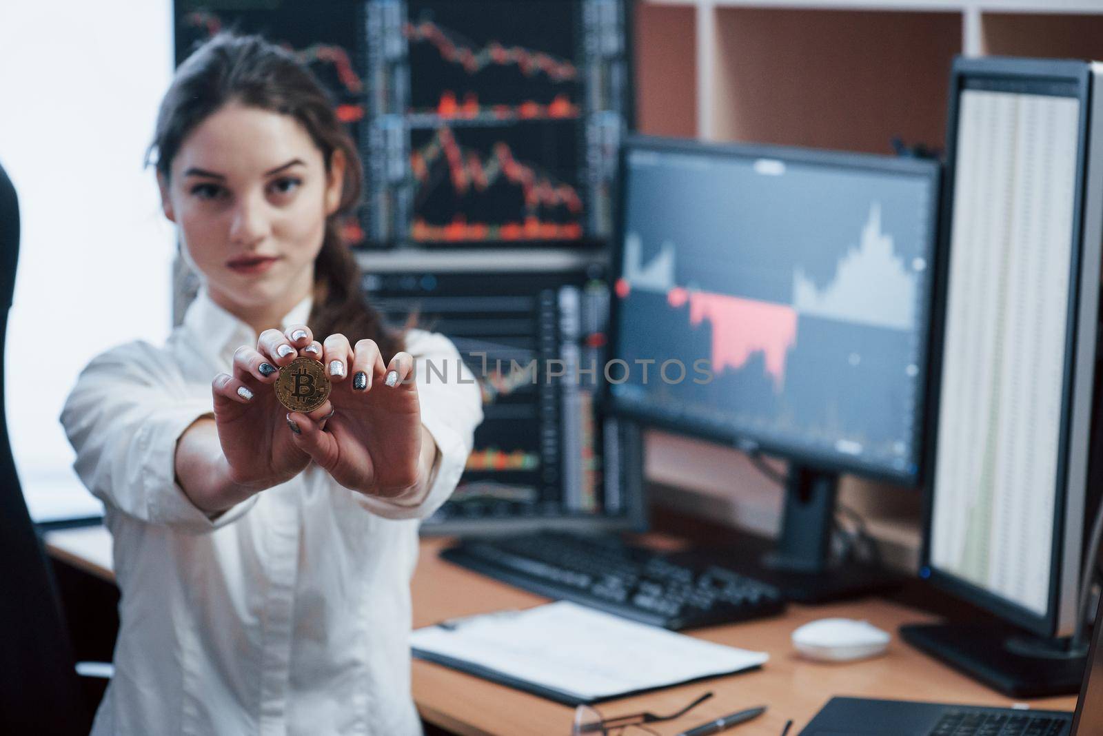 Modern currency. Beautiful woman holding bitcoin in hands while stands in modern office with many monitors with graphs.