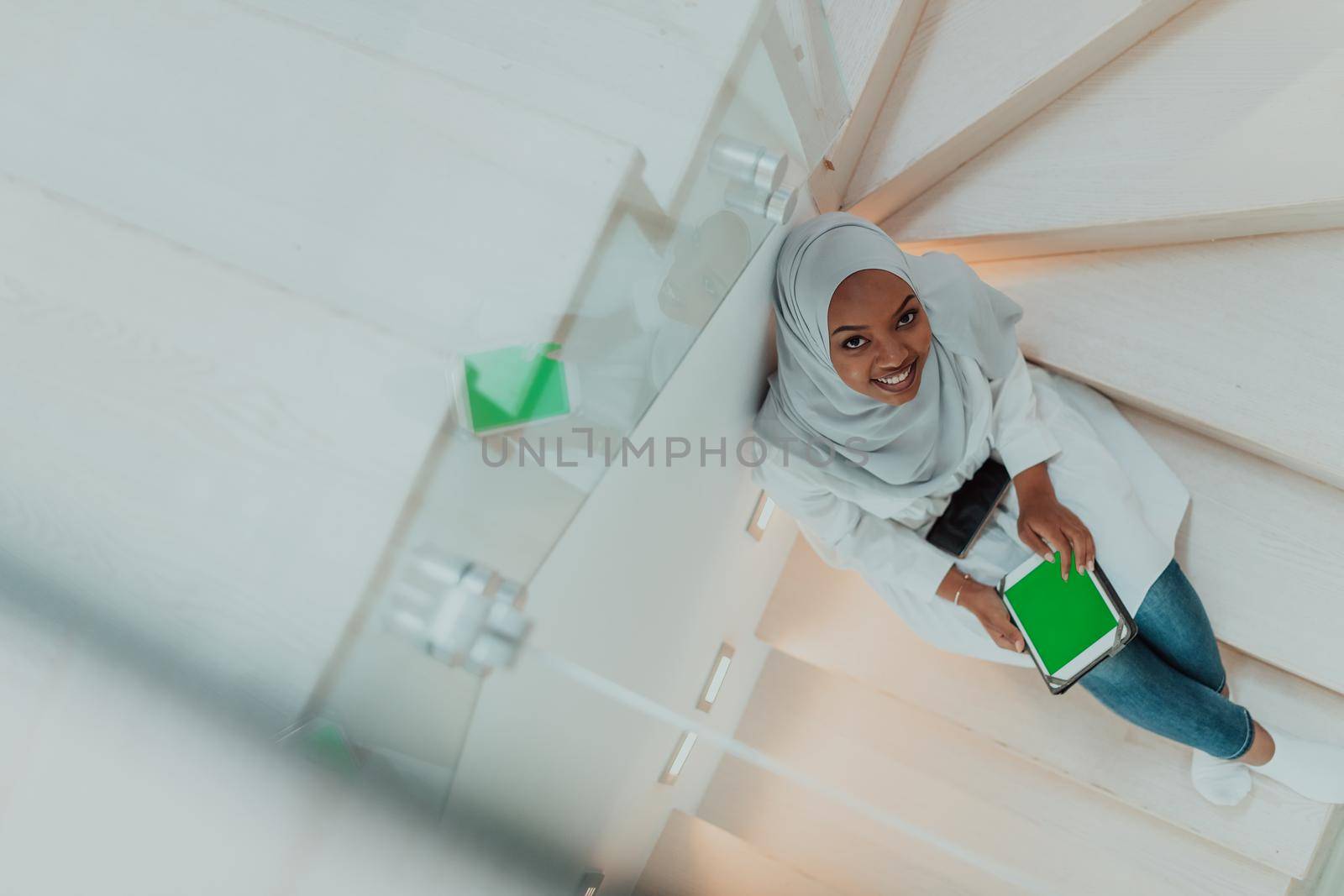 Young African modern Muslim woman using tablet computer while sitting on the stairs at home wearing hijab clothes top view. High-quality photo