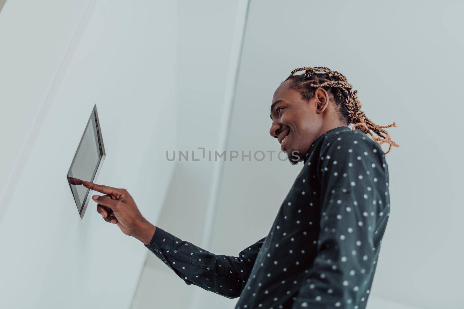 Smiling African American man using modern smart home system, controller on wall, positive young man switching temperature on thermostat or activating security alarm in apartment. High quality photo