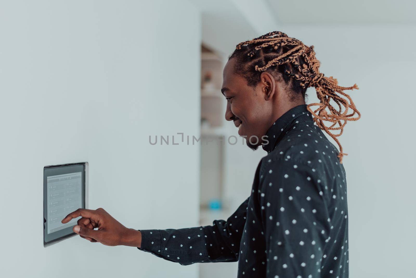 Smiling African American man using modern smart home system, controller on wall, positive young man switching temperature on thermostat or activating security alarm in apartment by dotshock