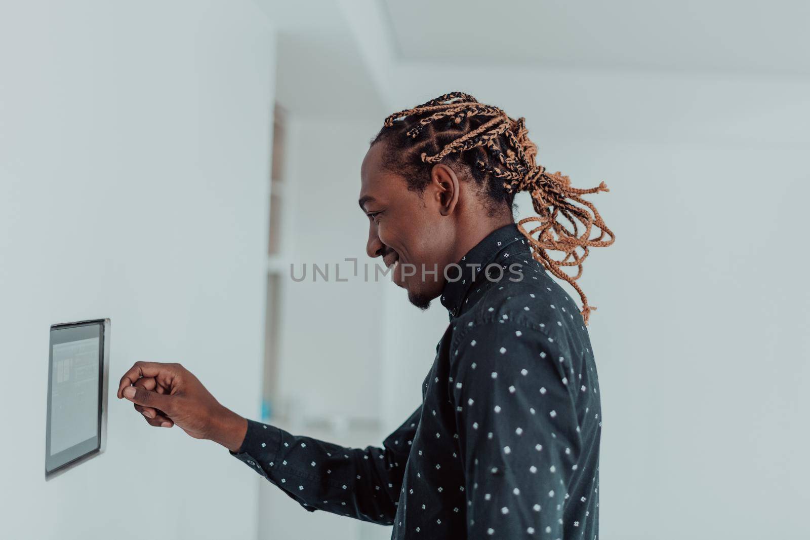 Smiling African American man using modern smart home system, controller on wall, positive young man switching temperature on thermostat or activating security alarm in apartment. High quality photo