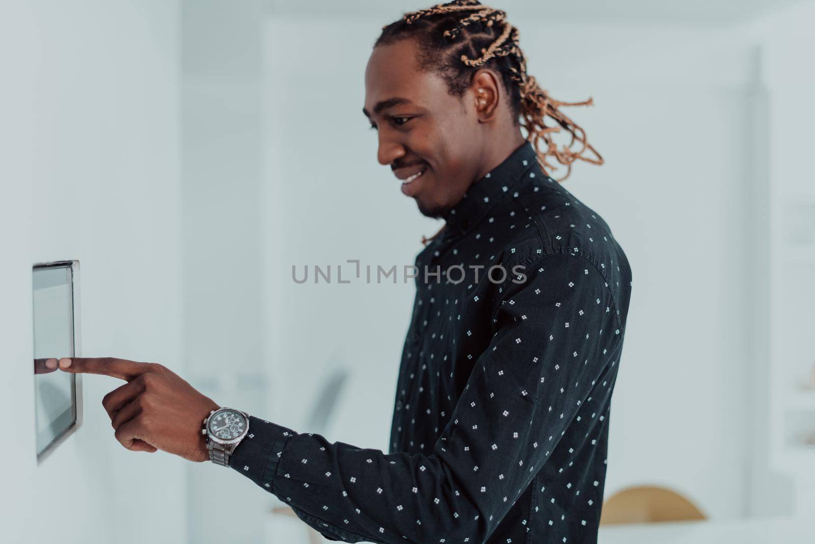 Smiling African American man using modern smart home system, controller on wall, positive young man switching temperature on thermostat or activating security alarm in apartment. High quality photo