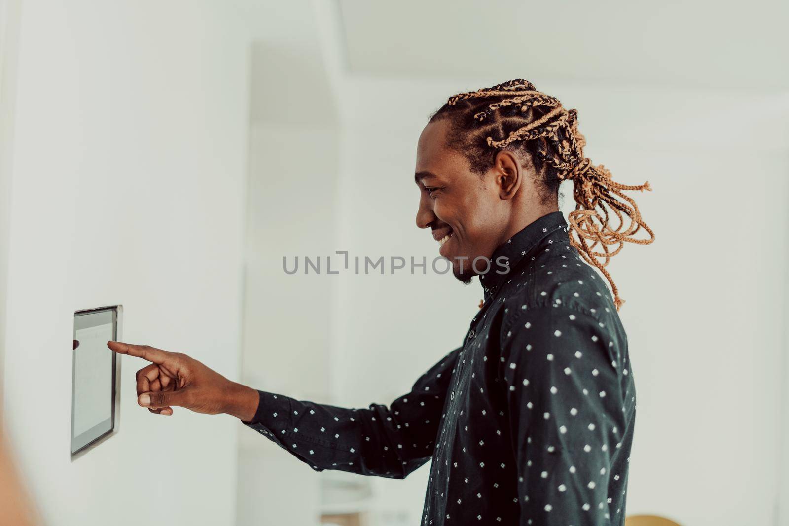 Smiling African American man using modern smart home system, controller on wall, positive young man switching temperature on thermostat or activating security alarm in apartment. High quality photo