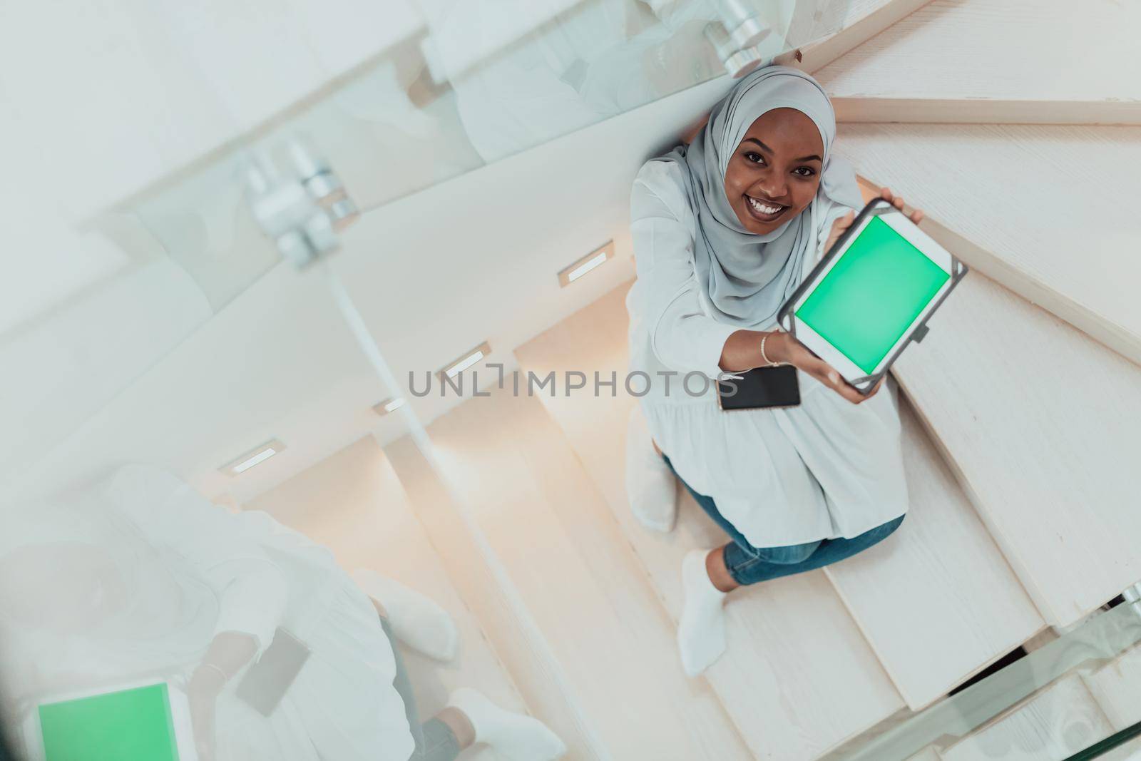 Young african modern muslim woman using tablet computer while sitting on the stairs at home wearing hijab clothes top view by dotshock