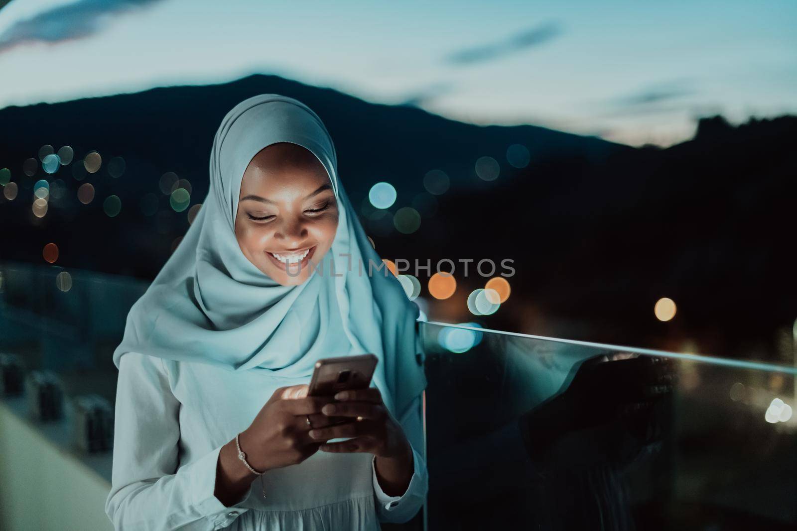 Young Muslim woman wearing scarf veil on urban city street at night texting on a smartphone with bokeh city light in the background. by dotshock