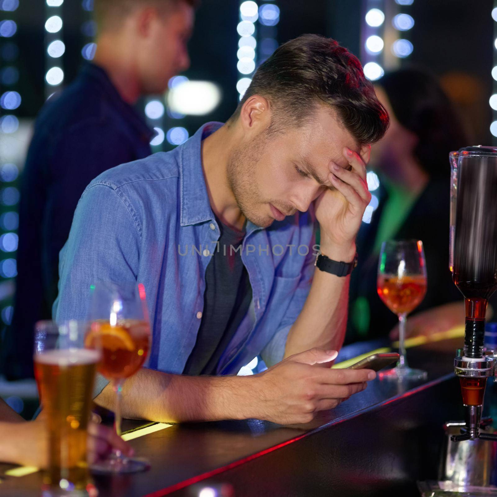 Shot of an upset looking young man reading a text on his cellphone while sitting at the counter of a nightclub.