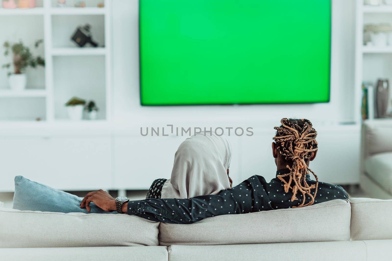 African Couple Sitting On Sofa Watching TV Together Chroma Green Screen Woman Wearing Islamic Hijab Clothes by dotshock