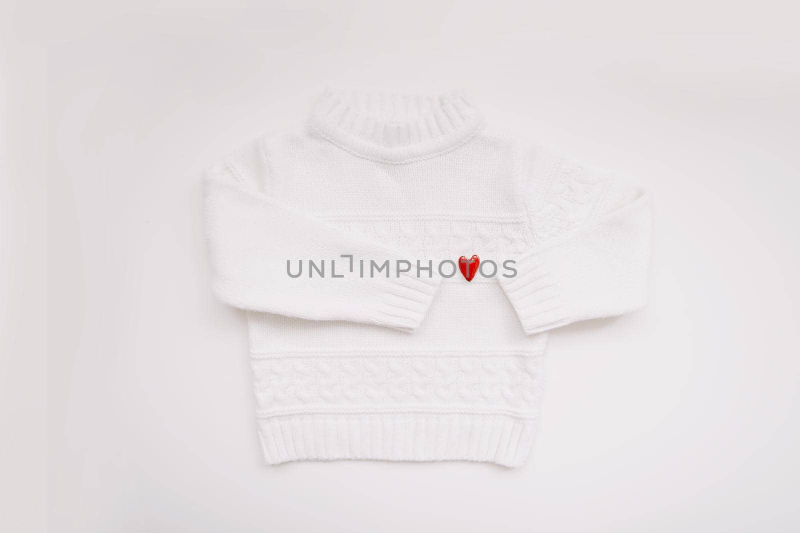 White woolen sweater with a red heart shaped pin on white background top view. Love, romance, fashion concept. Flat lay, copy space.