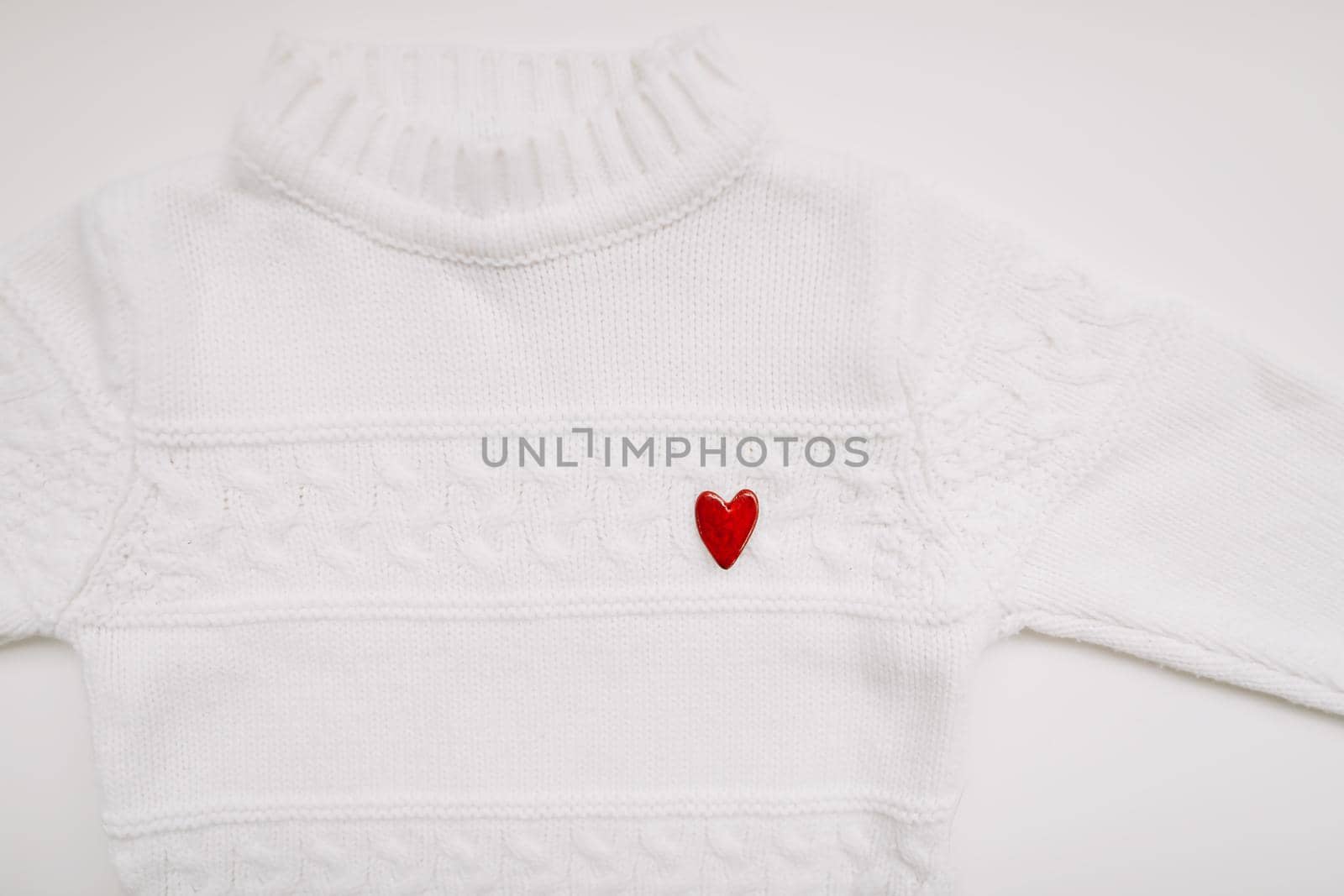 White woolen sweater with a red heart shaped pin on white background top view. Love, romance, fashion concept. Flat lay, copy space.