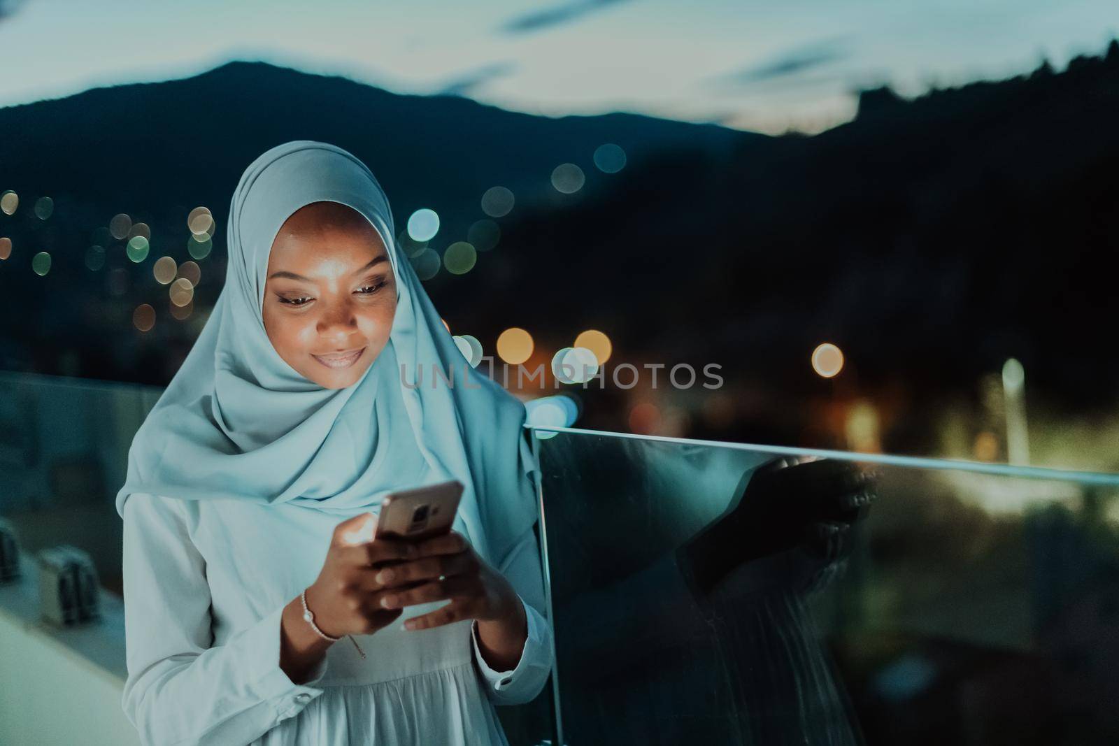 Young Muslim woman wearing scarf veil on urban city street at night texting on a smartphone with bokeh city light in the background. High-quality photo