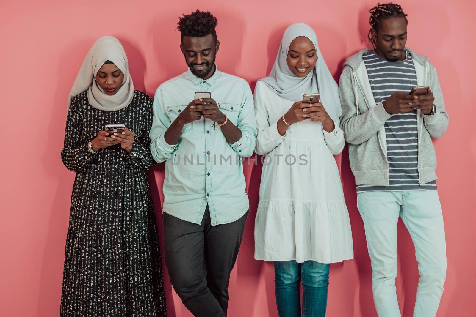 A group of African Muslim students use smartphones while standing in front of a pink background by dotshock