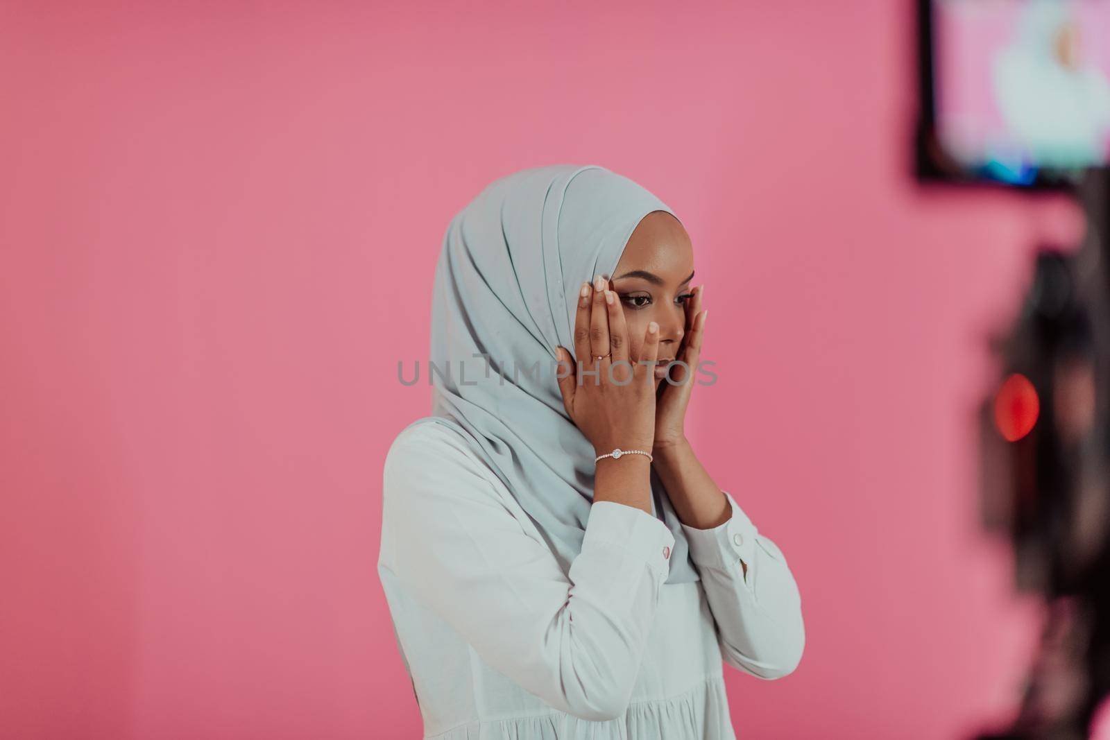 Modern African Muslim woman makes traditional prayer to God, keeps hands in praying gesture, wears traditional white clothes, has serious facial expression, isolated over plastic pink background by dotshock