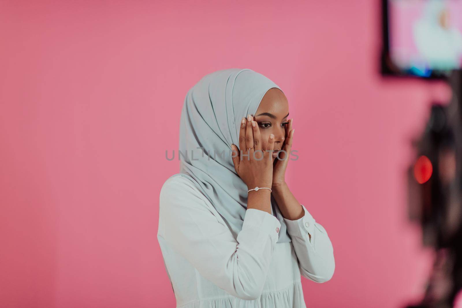 Modern African Muslim woman makes traditional prayer to God, keeps hands in praying gesture, wears traditional white clothes, has serious facial expression, isolated over plastic pink background. High-quality photo