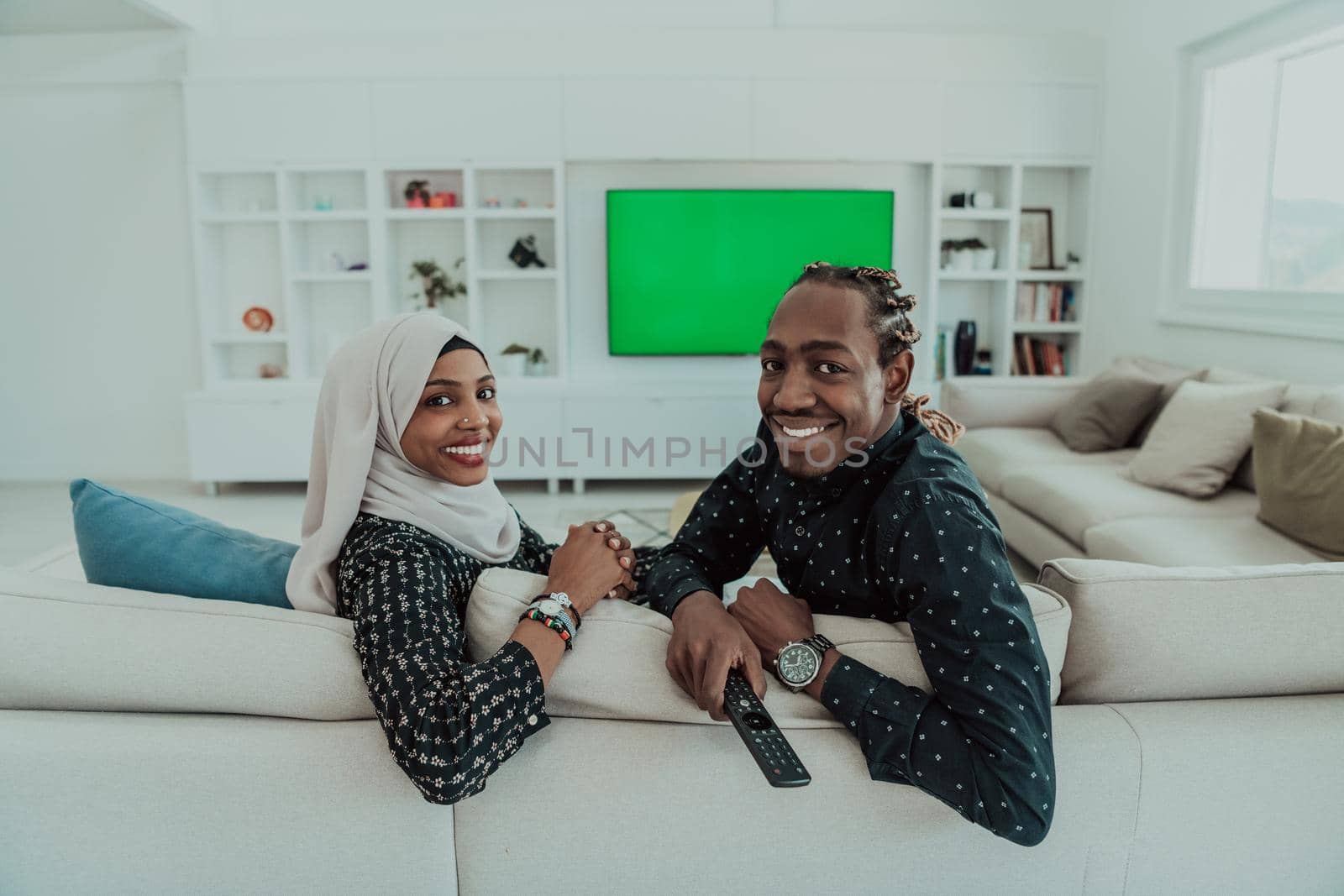 African Couple Sitting On Sofa Watching TV Together Chroma Green Screen Woman Wearing Islamic Hijab Clothes. High quality photo