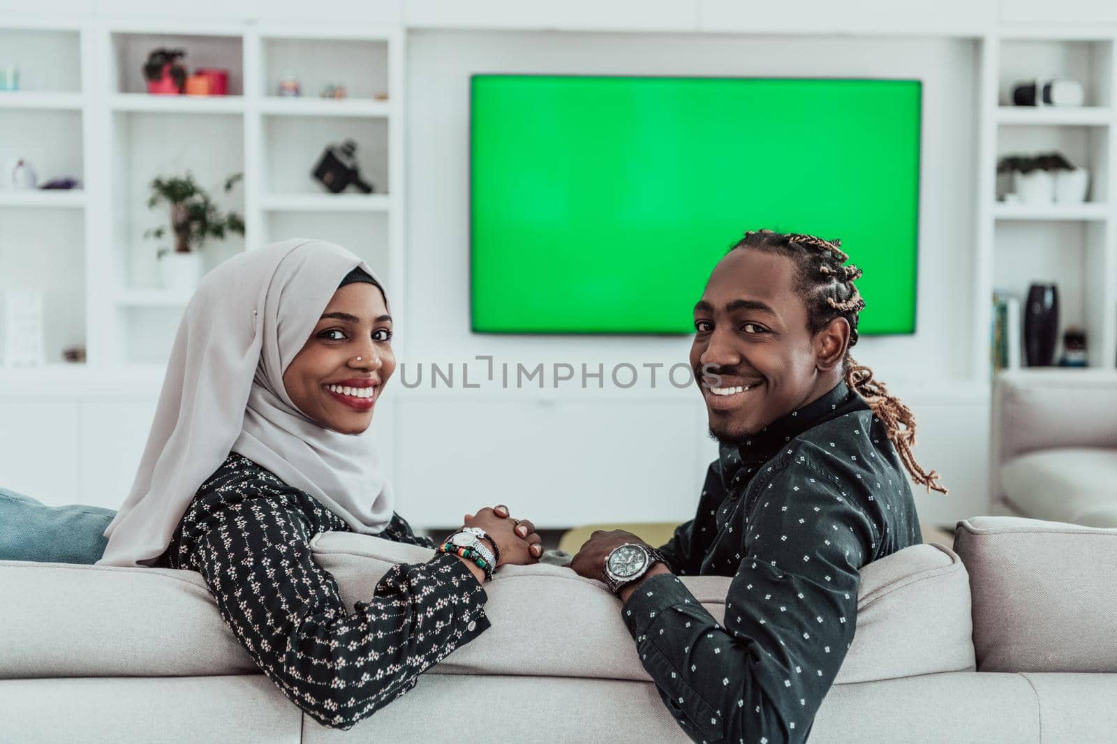 African Couple Sitting On Sofa Watching TV Together Chroma Green Screen Woman Wearing Islamic Hijab Clothes. High quality photo