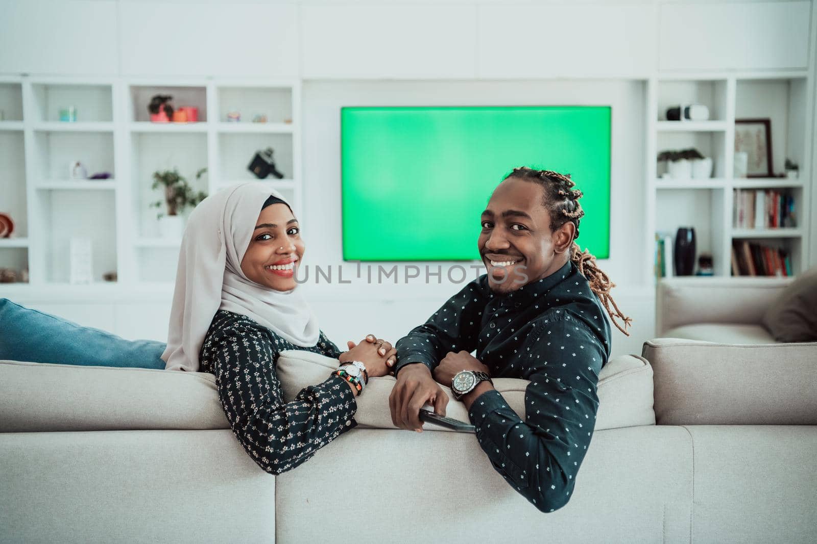 African Couple Sitting On Sofa Watching TV Together Chroma Green Screen Woman Wearing Islamic Hijab Clothes. High quality photo