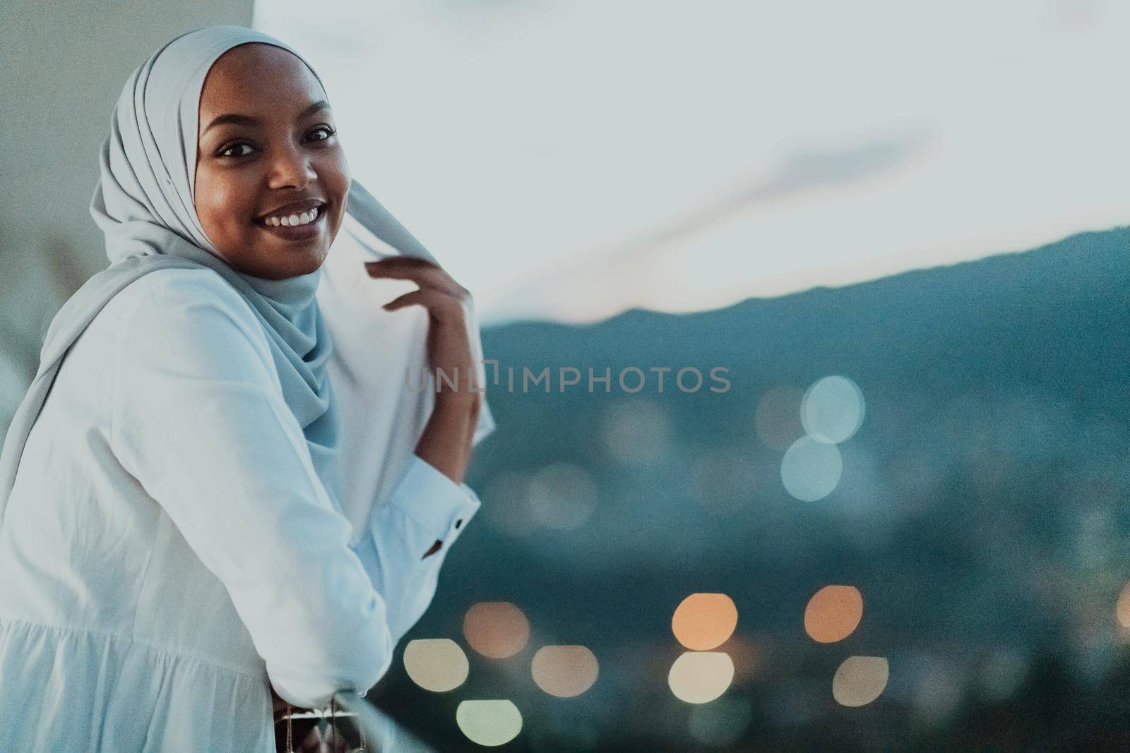 African Muslim woman in the night on a balcony smiling at the camera with city bokeh lights in the background. High-quality photo