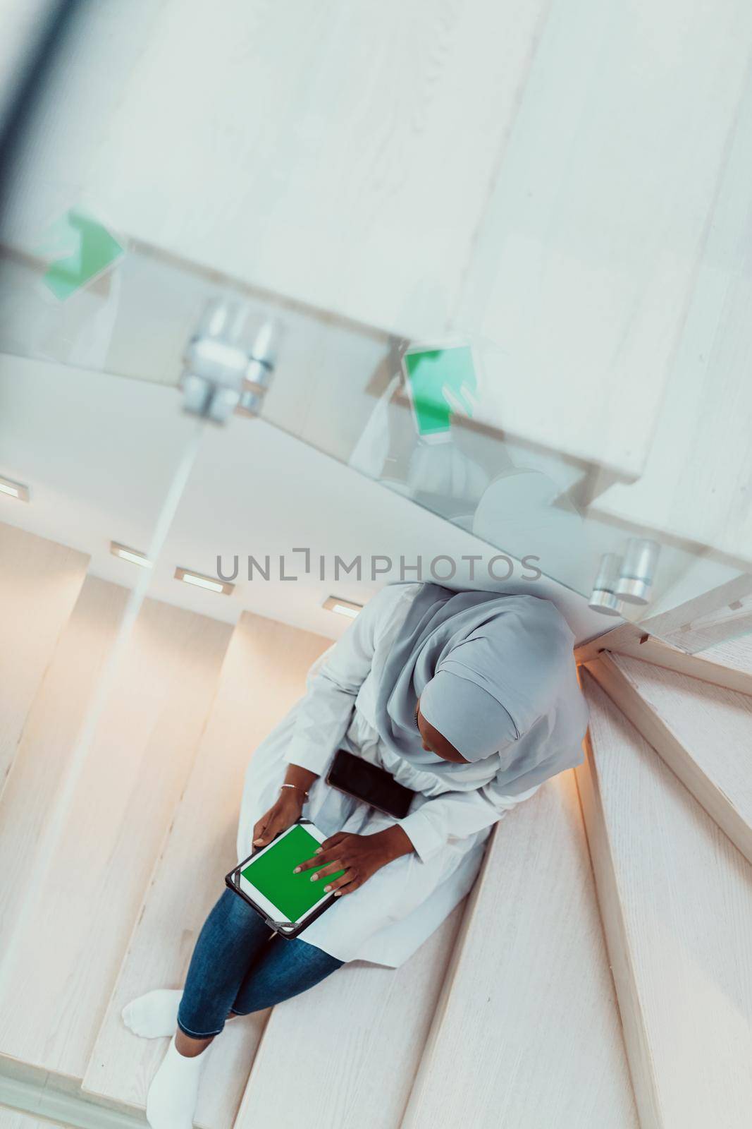 Young african modern muslim woman using tablet computer while sitting on the stairs at home wearing hijab clothes top view by dotshock