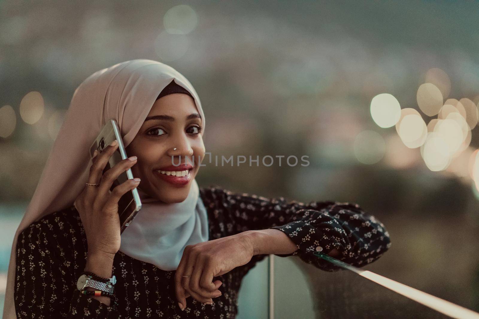 Young Muslim woman wearing scarf veil on urban city street at night texting on a smartphone with bokeh city light in the background. by dotshock
