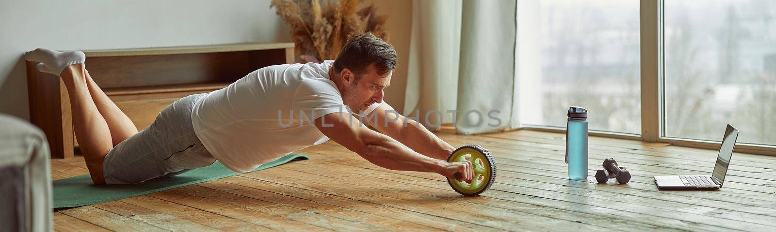 Athletic male is doing crunches with abs wheel while watching workout on notebook at home