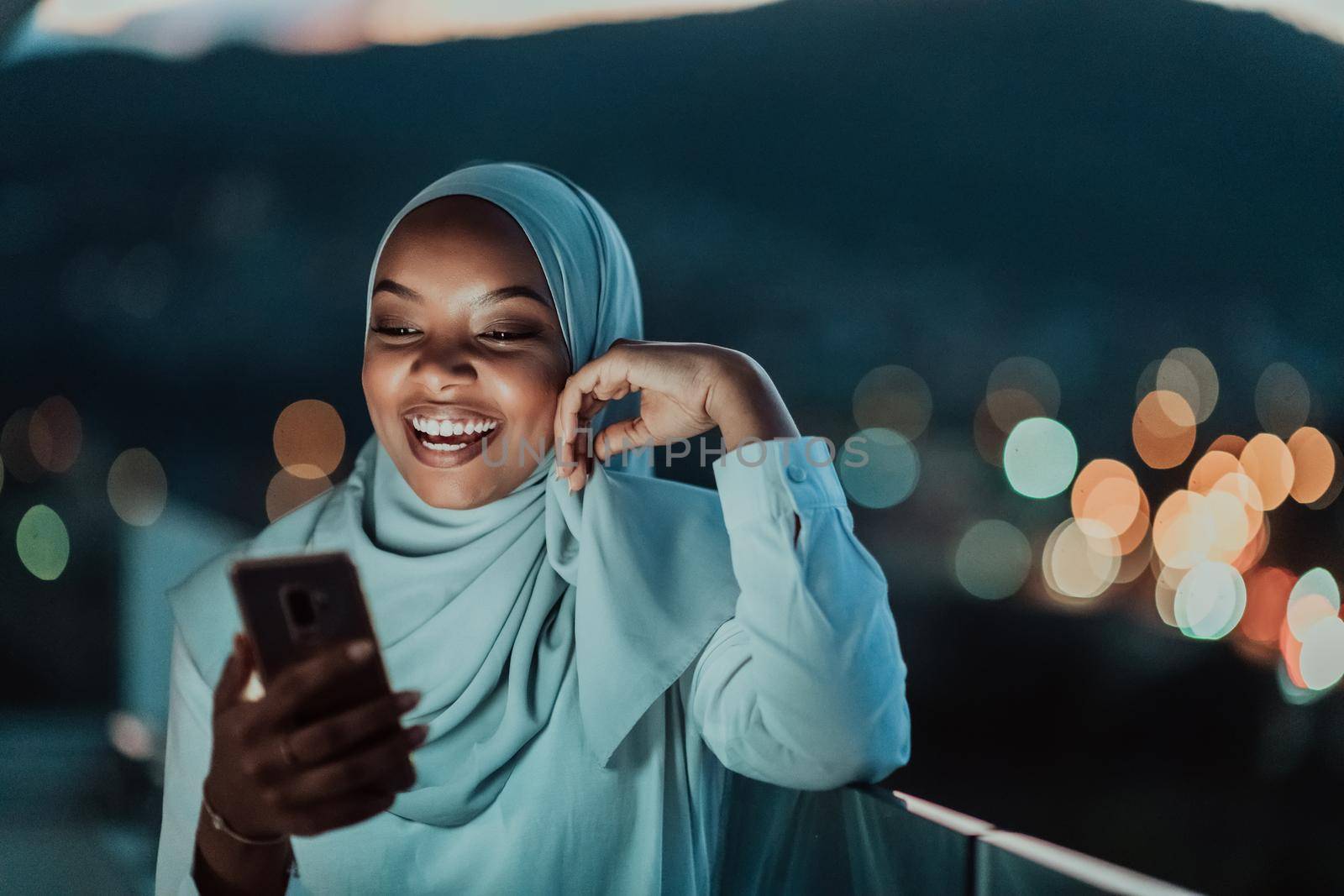 Young Muslim woman wearing scarf veil on urban city street at night texting on a smartphone with bokeh city light in the background. High-quality photo