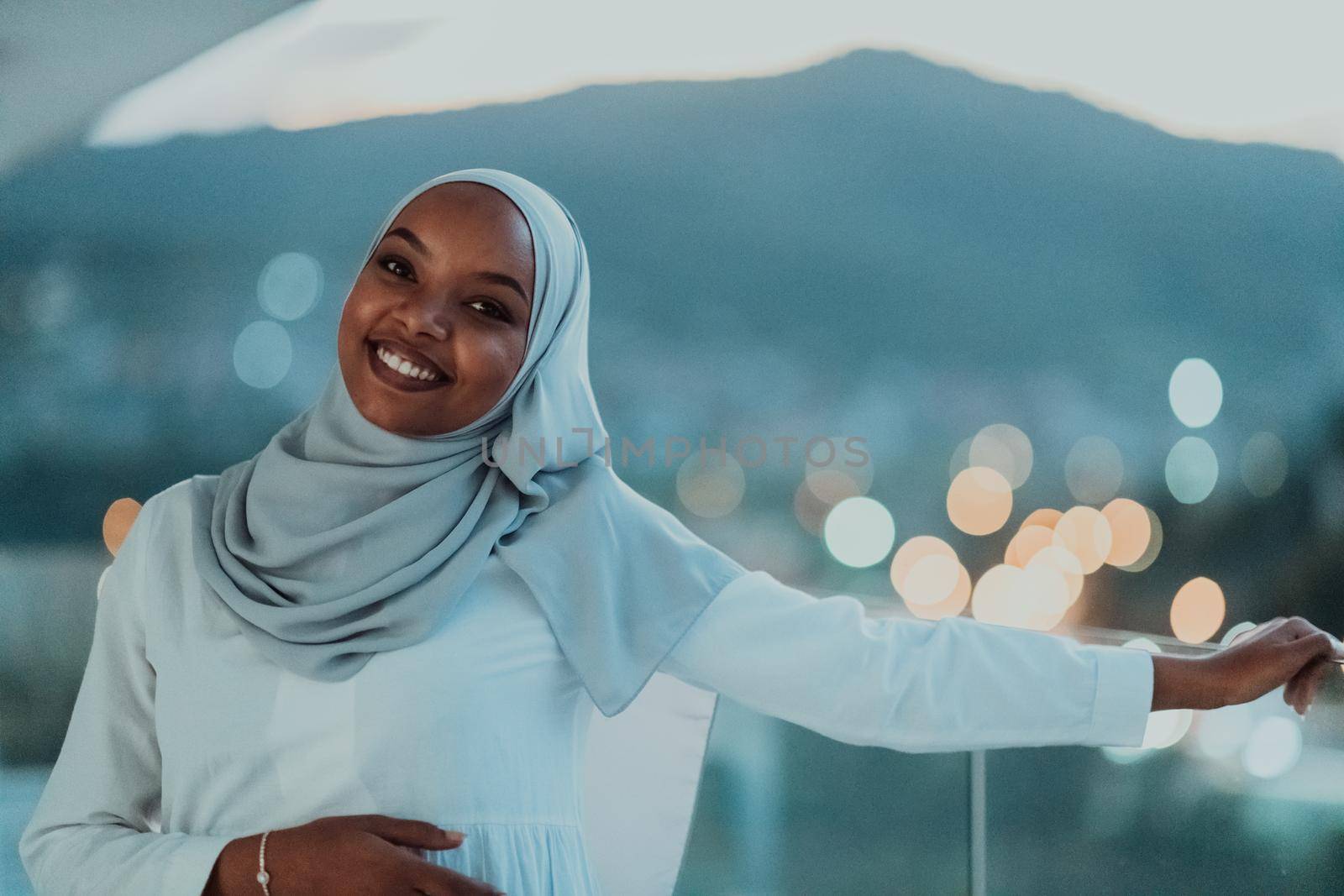 African Muslim woman in the night on a balcony smiling at the camera with city bokeh lights in the background. High-quality photo