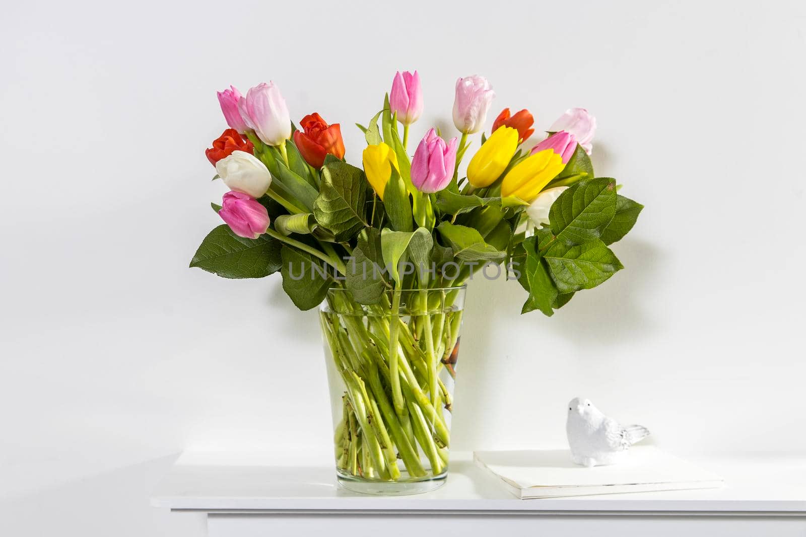 A bouquet of multi-colored tulips in a transparent vase and cup of tea on the white window.