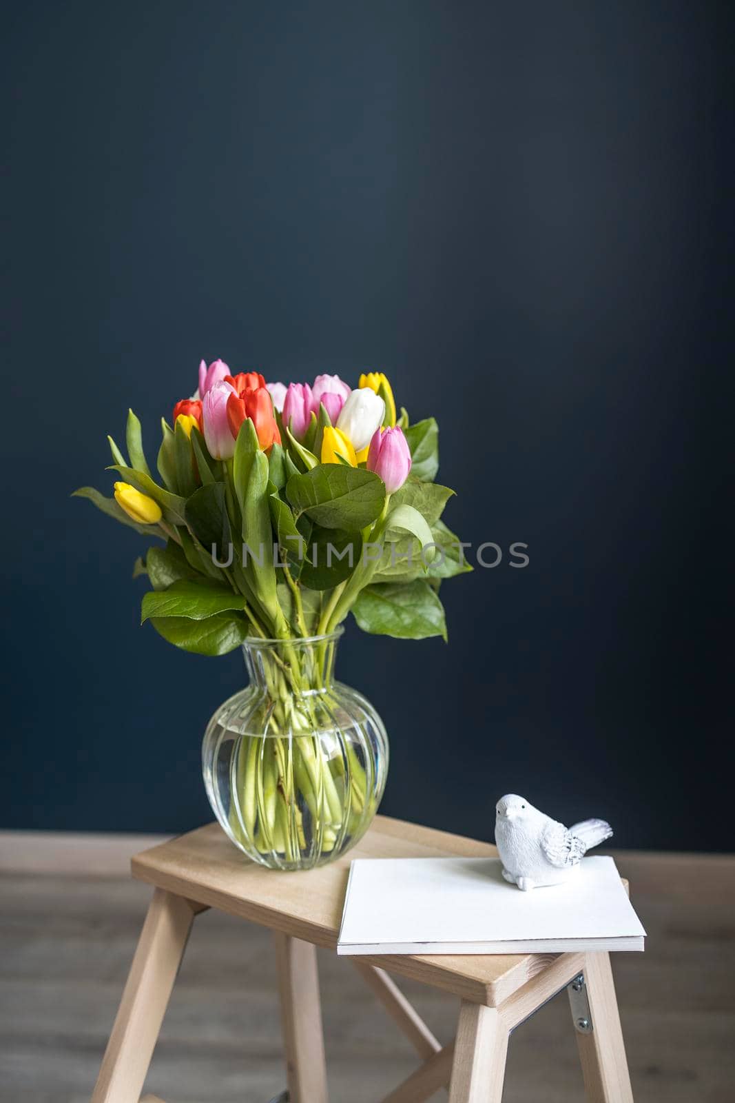 A bouquet of multi-colored tulips in a transparent vase on the tall chair with step against a dark blue background. White notebook.