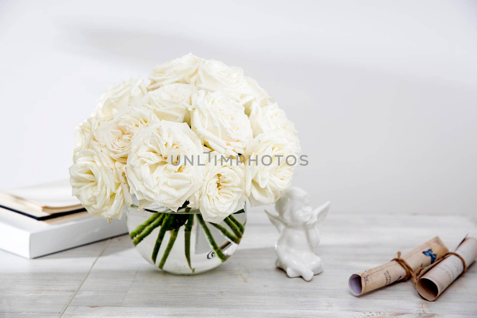 A bouquet of white roses in a round glass vase on a table with a figurine of angel and a book. by elenarostunova