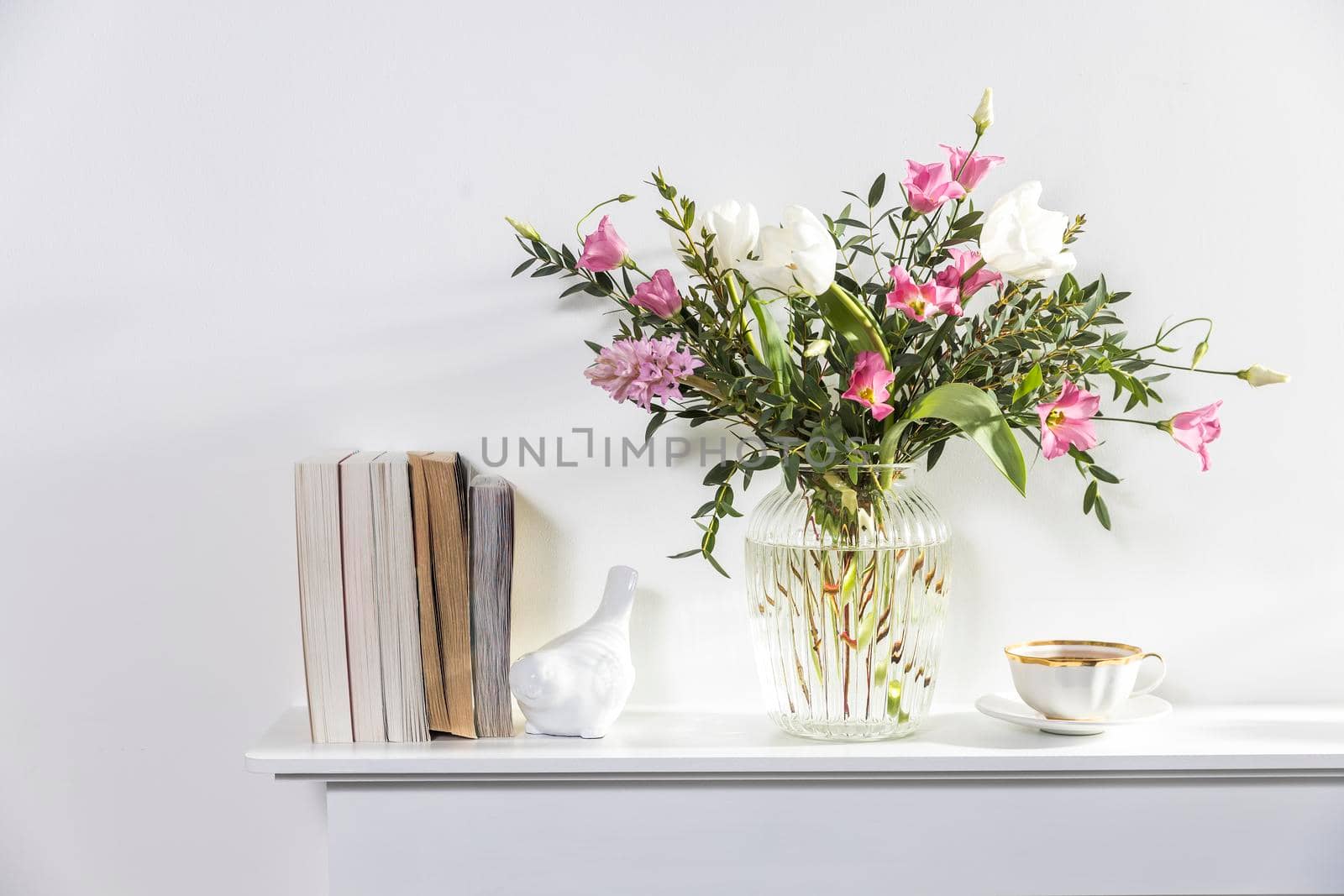 A minimalistic bouquet of white tulips, pink eustoma, hyacinth, eucalyptus in a fluted glass vase on a white panel of an artificial fireplace. by elenarostunova