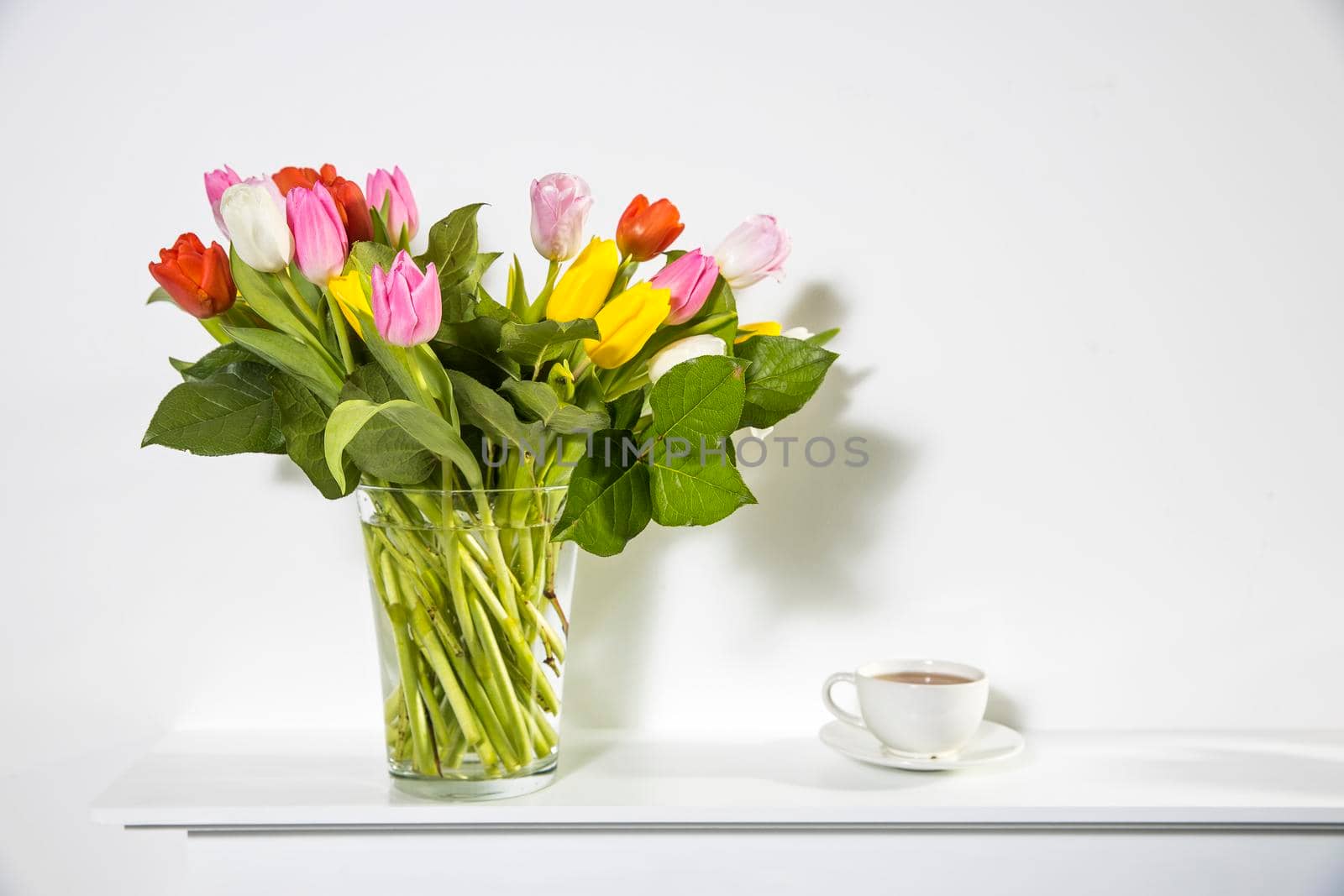 A bouquet of multi-colored tulips in a transparent vase and cup of tea on the white window.